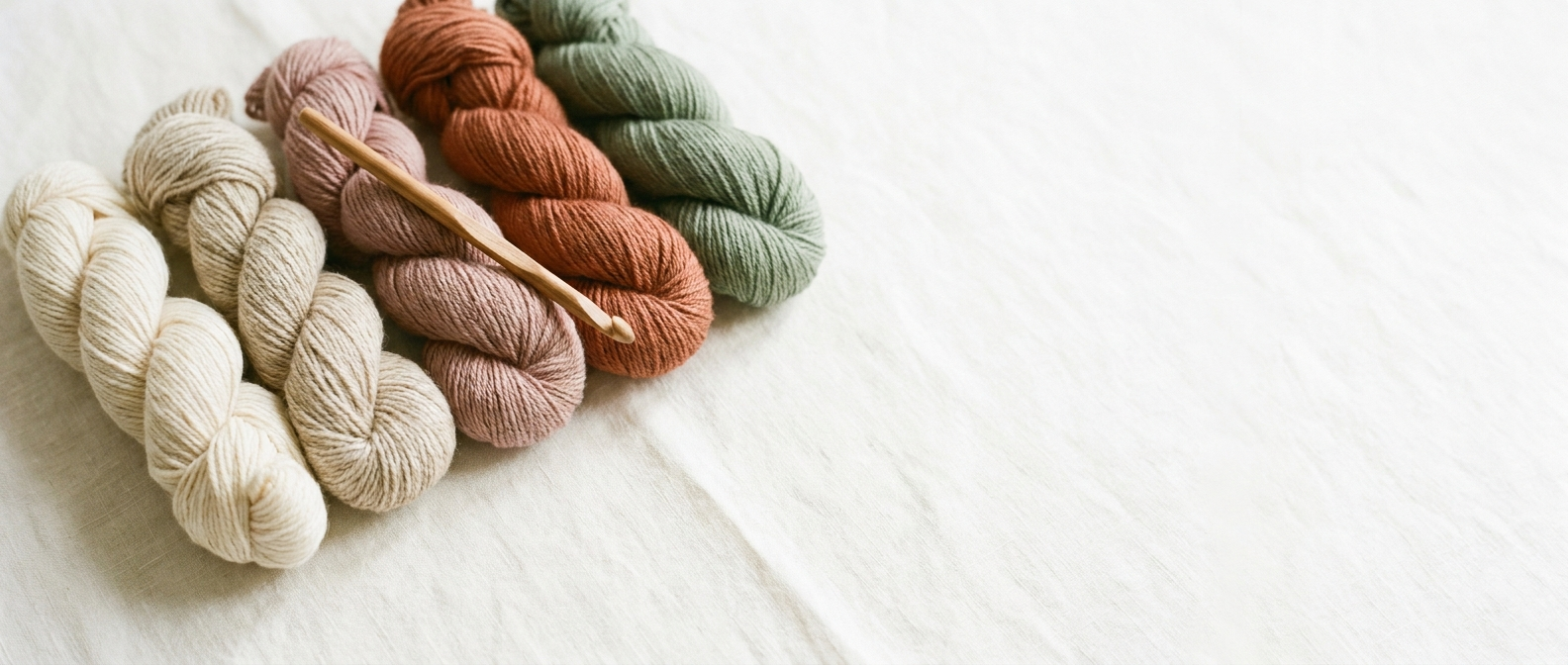 Yarn skeins in cream, dusty rose, terracotta and sage lined up on warm linen